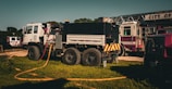 A fire truck and related emergency vehicles are parked on a grassy area, with hoses laid out for use. The fire truck is prominently displayed, featuring a large chassis and multiple compartments.