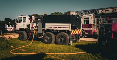 A fire truck and related emergency vehicles are parked on a grassy area, with hoses laid out for use. The fire truck is prominently displayed, featuring a large chassis and multiple compartments.
