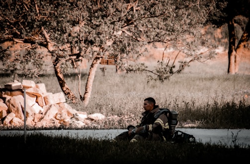 A person wearing a firefighter uniform sits on a grassy area near a pile of large stones. The setting appears to be outdoors, surrounded by trees and sparse vegetation. The lighting suggests it might be early morning or late afternoon, with warm, low-angle sunlight creating long shadows and a serene atmosphere.