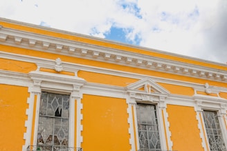 An architectural facade painted in bright yellow with intricate white detailing frames three tall windows. The windows feature decorative glass patterns, and the building's upper border includes ornate moldings. The scene is set against a partly cloudy blue sky.