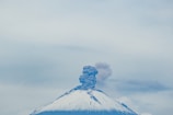 A snow-capped volcano with smoke and ash billowing from its summit under a cloudy sky.