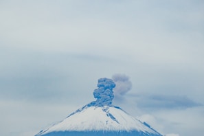 A snow-capped volcano with smoke and ash billowing from its summit under a cloudy sky.