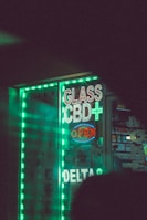 A storefront displays bright, neon signs that read 'Glass', 'CBD+', and 'Open', alongside other signage in a shop window. The signs are surrounded by glowing green strip lights, creating a vibrant and eye-catching effect. Inside the shop, various products are displayed, and there is a hint of colorful packaging. The overall scene is dimly lit, enhancing the prominence of the neon lights.