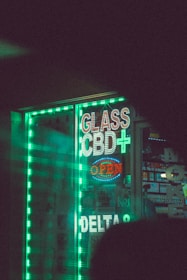 A storefront displays bright, neon signs that read 'Glass', 'CBD+', and 'Open', alongside other signage in a shop window. The signs are surrounded by glowing green strip lights, creating a vibrant and eye-catching effect. Inside the shop, various products are displayed, and there is a hint of colorful packaging. The overall scene is dimly lit, enhancing the prominence of the neon lights.