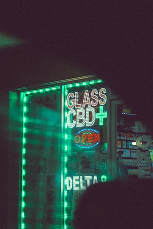 A storefront displays bright, neon signs that read 'Glass', 'CBD+', and 'Open', alongside other signage in a shop window. The signs are surrounded by glowing green strip lights, creating a vibrant and eye-catching effect. Inside the shop, various products are displayed, and there is a hint of colorful packaging. The overall scene is dimly lit, enhancing the prominence of the neon lights.