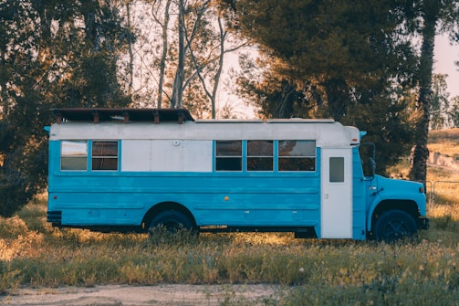 A bright blue and white bus is parked in a grassy area, surrounded by tall trees. The bus is customized, with a panel on the roof that could serve as a solar panel or storage. The setting is tranquil, with the sunlight filtering through the trees, creating a peaceful outdoor scene.