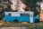 A bright blue and white bus is parked in a grassy area, surrounded by tall trees. The bus is customized, with a panel on the roof that could serve as a solar panel or storage. The setting is tranquil, with the sunlight filtering through the trees, creating a peaceful outdoor scene.