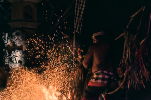 A person wearing traditional attire appears to be involved in a ritual or performance involving fire. Sparks fly as the flames rise against the dark background, adding a dynamic and mystical quality to the scene. Ornate stone carvings are visible in the dimly lit surroundings, hinting at a cultural or religious setting. The motion of the sparks and the posture of the person suggest a moment of intense focus and activity.