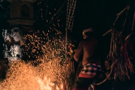 A person wearing traditional attire appears to be involved in a ritual or performance involving fire. Sparks fly as the flames rise against the dark background, adding a dynamic and mystical quality to the scene. Ornate stone carvings are visible in the dimly lit surroundings, hinting at a cultural or religious setting. The motion of the sparks and the posture of the person suggest a moment of intense focus and activity.
