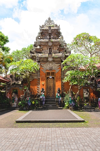 An ornate, traditional Balinese temple gate with intricate carvings and decorations. The structure features a prominent central doorway and is flanked by statues. Surrounding the temple are lush tropical plants and trees under a partly cloudy sky.