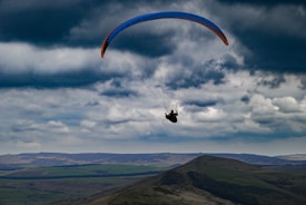 A paraglider is soaring over a wide expanse of green hills under a cloudy sky. The landscape features gently rolling terrain with varying shades of green and blue, blending into the distant horizon.