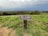A wooden sign is posted on a grassy hill, stating 'Area Closed for Resource Protection.' The landscape in the background includes dense green vegetation and stretches into a panoramic view of a town or city below, beneath overcast skies.