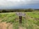 A wooden sign is posted on a grassy hill, stating 'Area Closed for Resource Protection.' The landscape in the background includes dense green vegetation and stretches into a panoramic view of a town or city below, beneath overcast skies.