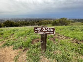 A wooden sign is posted on a grassy hill, stating 'Area Closed for Resource Protection.' The landscape in the background includes dense green vegetation and stretches into a panoramic view of a town or city below, beneath overcast skies.