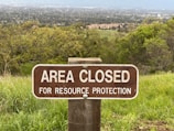A wooden sign positioned in a grassy area reads 'Area Closed for Resource Protection.' The background features a wide view of a landscape, including trees and what appears to be a town or settlement further away.