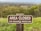 A wooden sign positioned in a grassy area reads 'Area Closed for Resource Protection.' The background features a wide view of a landscape, including trees and what appears to be a town or settlement further away.