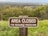 A wooden sign positioned in a grassy area reads 'Area Closed for Resource Protection.' The background features a wide view of a landscape, including trees and what appears to be a town or settlement further away.