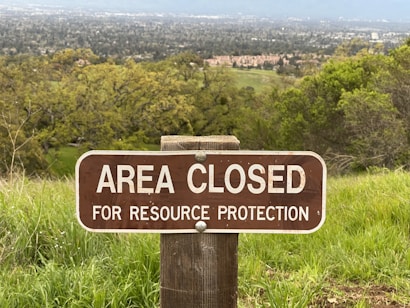 A wooden sign positioned in a grassy area reads 'Area Closed for Resource Protection.' The background features a wide view of a landscape, including trees and what appears to be a town or settlement further away.