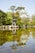 A well-manicured garden with a reflective pond in the foreground, featuring a traditional Japanese pagoda and a stone lantern. Lush greenery and trees surround the area, and a large, historic Japanese castle is prominently visible in the background.
