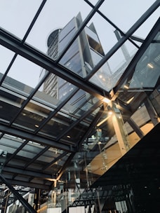 A modern architectural scene features a tall skyscraper with a glass facade, viewed through an overhanging glass and steel structure that reflects and refracts light. The intricate framework creates a dynamic and futuristic atmosphere.