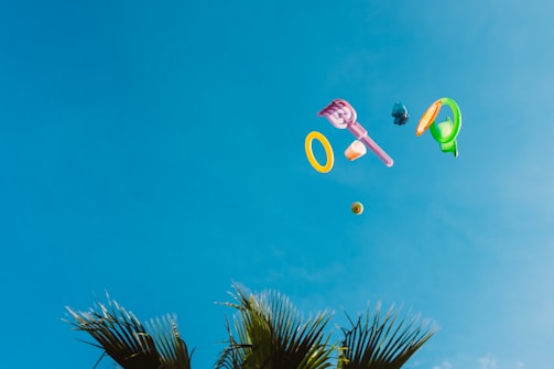 Various colorful plastic toys appear to be floating or thrown into a clear blue sky. Palm fronds are visible at the bottom of the image, suggesting a tropical or outdoor setting.