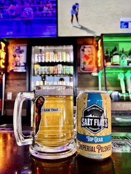A glass mug filled with beer sits next to a can labeled 'Salt Flats Brewing Co. Top Gear Imperial Pilsner' on a bar counter. In the background are colorful shelves stocked with various liquor bottles and two televisions, one showing a sports event.