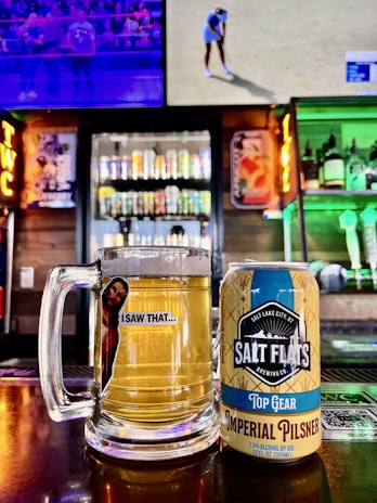 A glass mug filled with beer sits next to a can labeled 'Salt Flats Brewing Co. Top Gear Imperial Pilsner' on a bar counter. In the background are colorful shelves stocked with various liquor bottles and two televisions, one showing a sports event.