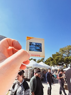 A hand holding a vintage Kodak slide with an image of a car and building is prominently featured against a clear blue sky. In the background, several people walk among market stalls with white canopies. The scene suggests a lively outdoor market setting with trees and a sunny atmosphere.