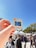 A hand holding a vintage Kodak slide with an image of a car and building is prominently featured against a clear blue sky. In the background, several people walk among market stalls with white canopies. The scene suggests a lively outdoor market setting with trees and a sunny atmosphere.