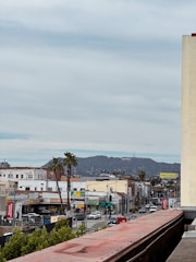 A cityscape featuring a street lined with various businesses and vehicles. Palm trees dot the scene, and in the distance, the iconic Hollywood sign is visible on a hillside. Buildings display colorful signage and advertisements, adding to the urban atmosphere.