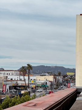 A cityscape featuring a street lined with various businesses and vehicles. Palm trees dot the scene, and in the distance, the iconic Hollywood sign is visible on a hillside. Buildings display colorful signage and advertisements, adding to the urban atmosphere.