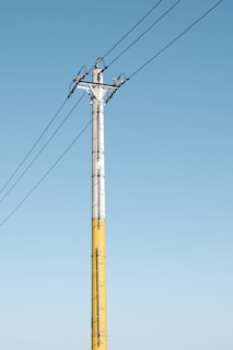 A tall utility pole is visible against a clear blue sky. The pole, painted in yellow and gray, supports several wires extending across the image. Metal brackets and clamps secure the wires to the pole.