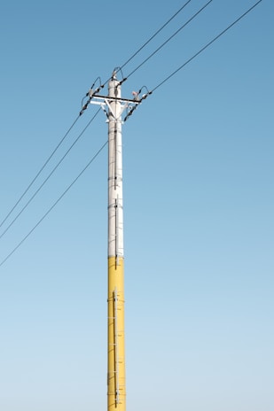 A tall utility pole is visible against a clear blue sky. The pole, painted in yellow and gray, supports several wires extending across the image. Metal brackets and clamps secure the wires to the pole.