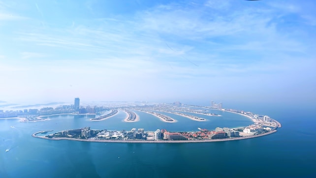 An aerial view of a man-made island resembling a palm tree, set in a bright blue sea. The island is densely populated with buildings, featuring several long, slender fronds extending into the water. In the background, a hazy skyline of tall buildings rises against a slightly cloudy, yet bright blue sky.