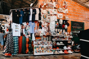 A vibrant market stall displaying a wide variety of colorful merchandise, including t-shirts with diverse prints, patterned fabric, handbags with creative designs, hats of different styles and colors, and an array of small figurines and souvenirs. The brick wall background adds to the rustic charm of the setting, while a bicycle partially visible on the side hints at the bustling activity around.