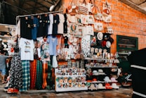 A vibrant market stall displaying a wide variety of colorful merchandise, including t-shirts with diverse prints, patterned fabric, handbags with creative designs, hats of different styles and colors, and an array of small figurines and souvenirs. The brick wall background adds to the rustic charm of the setting, while a bicycle partially visible on the side hints at the bustling activity around.