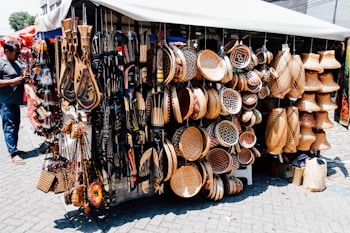 A market stall displays a diverse range of handcrafted items including intricately patterned baskets, wooden carvings, and decorative wall hangings. The items are artfully arranged, showing a variety of textures and designs. A person stands to the left, possibly examining or purchasing items.