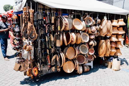 A market stall displays a diverse range of handcrafted items including intricately patterned baskets, wooden carvings, and decorative wall hangings. The items are artfully arranged, showing a variety of textures and designs. A person stands to the left, possibly examining or purchasing items.