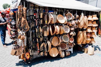A market stall displays a diverse range of handcrafted items including intricately patterned baskets, wooden carvings, and decorative wall hangings. The items are artfully arranged, showing a variety of textures and designs. A person stands to the left, possibly examining or purchasing items.