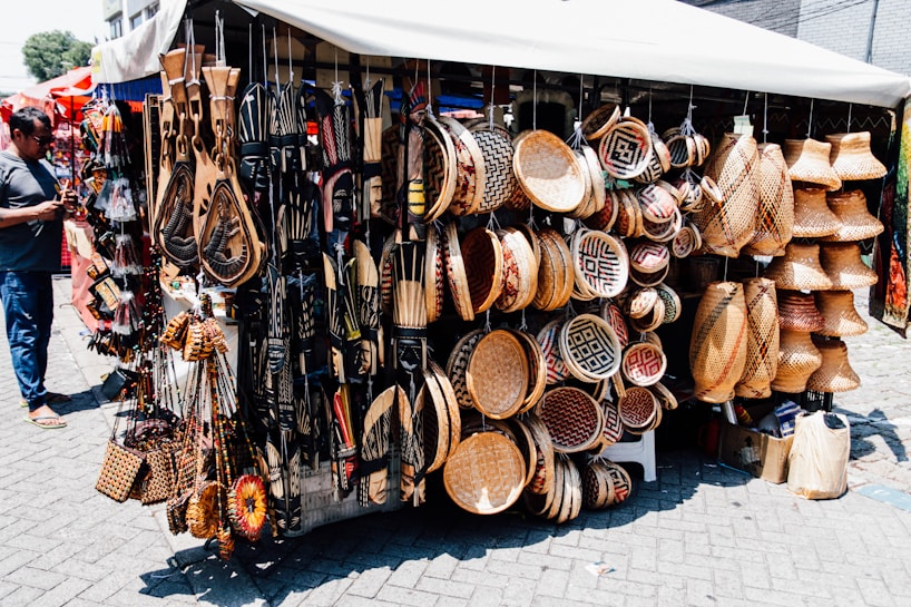 A market stall displays a diverse range of handcrafted items including intricately patterned baskets, wooden carvings, and decorative wall hangings. The items are artfully arranged, showing a variety of textures and designs. A person stands to the left, possibly examining or purchasing items.