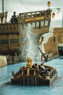 A large wooden ship with the name 'Santa Marian' is prominently featured, docked in a water-filled amusement park attraction. In the foreground, a small boat resembling a ride vehicle is carrying passengers, and there is a splash of water indicating movement or a ride element. The setting appears to be themed with historical or pirate-like elements.