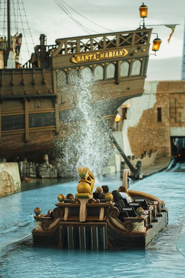 A large wooden ship with the name 'Santa Marian' is prominently featured, docked in a water-filled amusement park attraction. In the foreground, a small boat resembling a ride vehicle is carrying passengers, and there is a splash of water indicating movement or a ride element. The setting appears to be themed with historical or pirate-like elements.