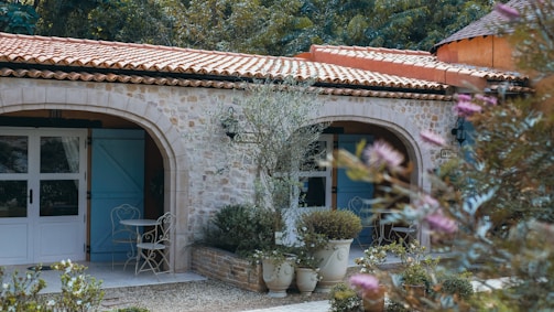A Mediterranean-style building with stone walls features blue shutters and a terra cotta tiled roof. In front of the building, there is a small patio area with wrought iron furniture. Several potted plants and trees add greenery to the scene, and there are decorative elements like lanterns on the walls.