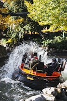 A river raft ride splashes through water surrounded by lush greenery. The bright orange raft is filled with passengers, some wearing protective gear. Water sprays dramatically around the vessel, indicating high-speed movement and excitement.