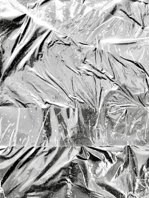Crinkled and reflective surface that resembles aluminum foil, with a textured appearance characterized by folds, creases, and shiny highlights. The light reflects off various angles, creating a dynamic pattern.