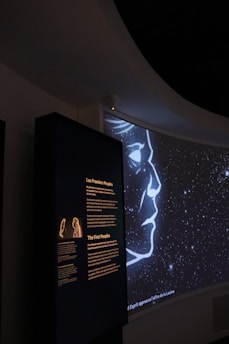 A dark exhibition space featuring an illuminated panel with text in both French and English, discussing the 'First Peoples.' Adjacent is a projection showing a stylized outline of a face amidst a starry night background.