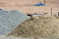 A large pile of sand and another pile of gravel are visible in an outdoor construction site. The background is a barren stretch of land with blurred objects and what appears to be construction debris.
