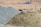 A large pile of sand and another pile of gravel are visible in an outdoor construction site. The background is a barren stretch of land with blurred objects and what appears to be construction debris.