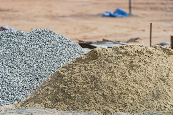 A large pile of sand and another pile of gravel are visible in an outdoor construction site. The background is a barren stretch of land with blurred objects and what appears to be construction debris.