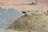 A large pile of sand and another pile of gravel are visible in an outdoor construction site. The background is a barren stretch of land with blurred objects and what appears to be construction debris.